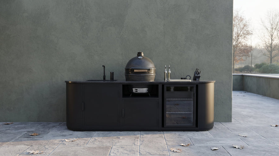 Zwarte buitenkeuken met kamado, spoelbak en koelkast tegen een muur op een terras in de herfst